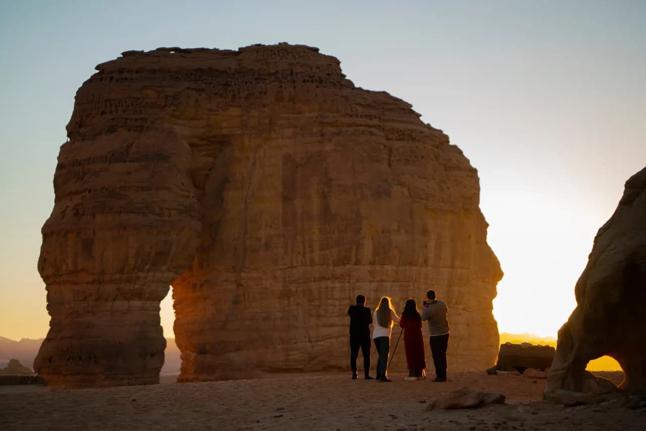 Elephant Rock silhouetted against a sunset sky in AlUla, with visitors visible at its base for scale