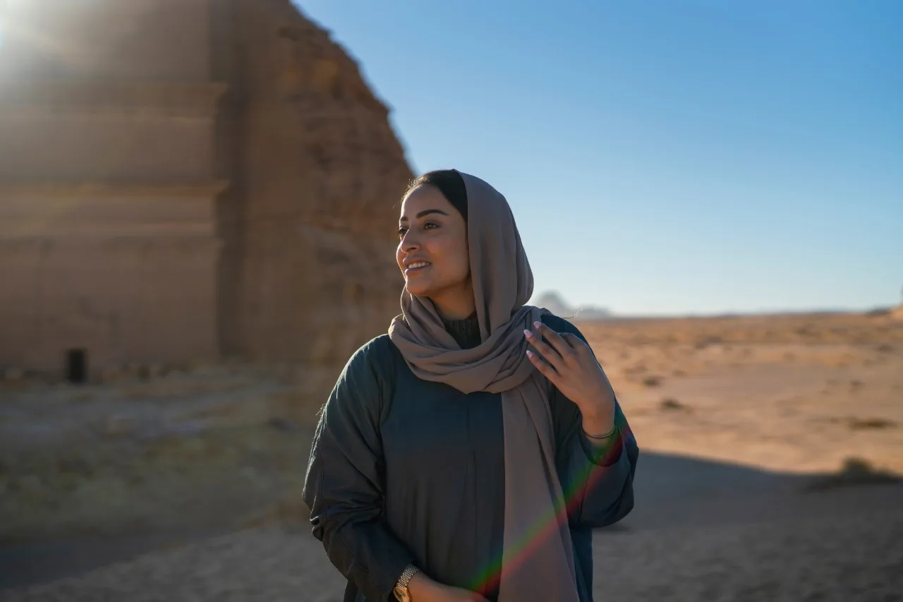 A lone visitor in traditional dress standing before a monumental Hegra tomb, emphasising the sense of solitude and scale